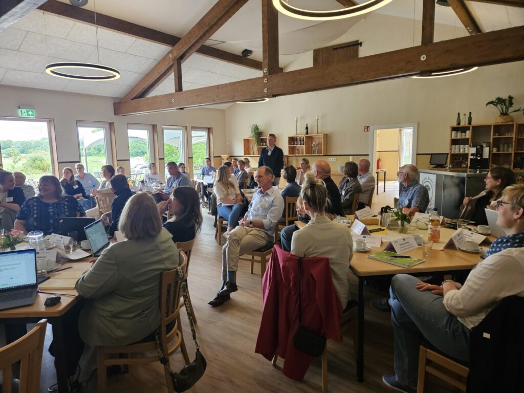 Viele Menschen sitzen auf Stühlen um Tische und schauen nach hinten zu einem Mann, der steht