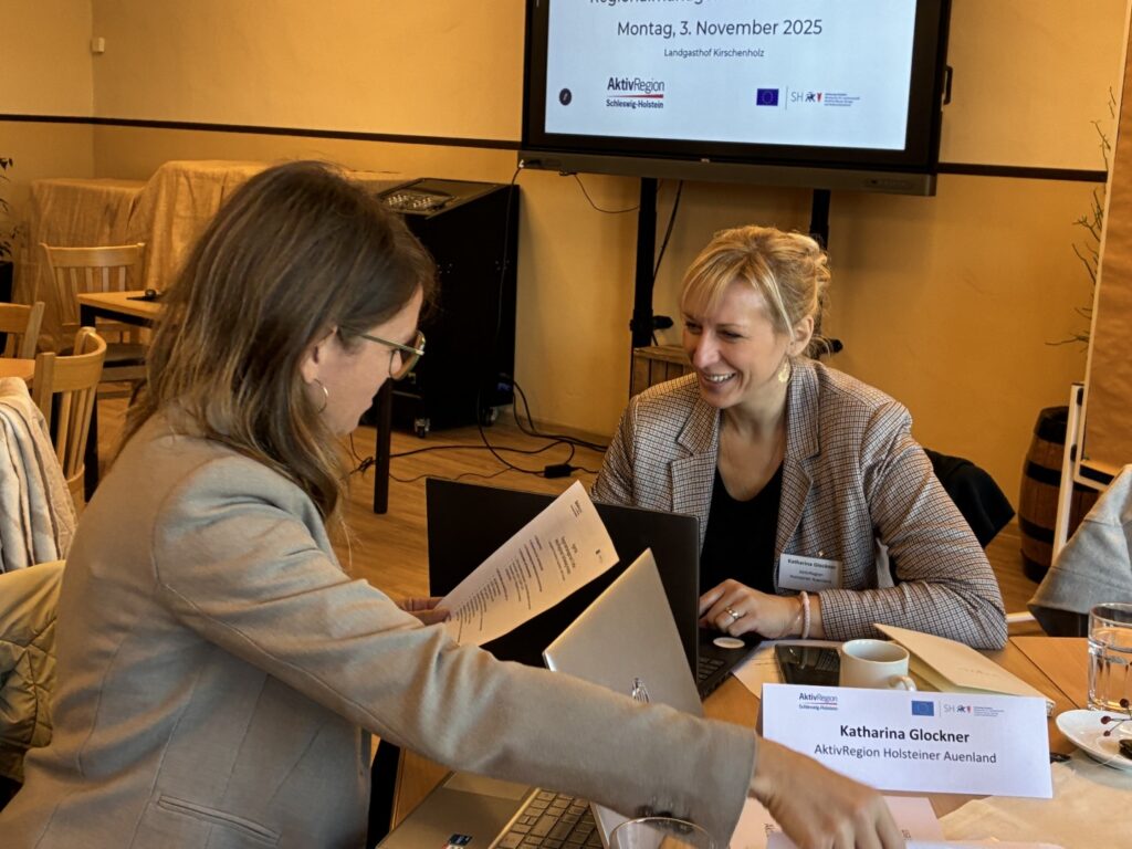 Zwei Frauen sitzen an einem Tisch und besprechen etwas. eine Frau lächelt, die andere hält ein Papier in der Hand Zwei Frauen sitzen an einem Tisch und besprechen etwas. eine Frau lächelt, die andere hält ein Papier in der Hand