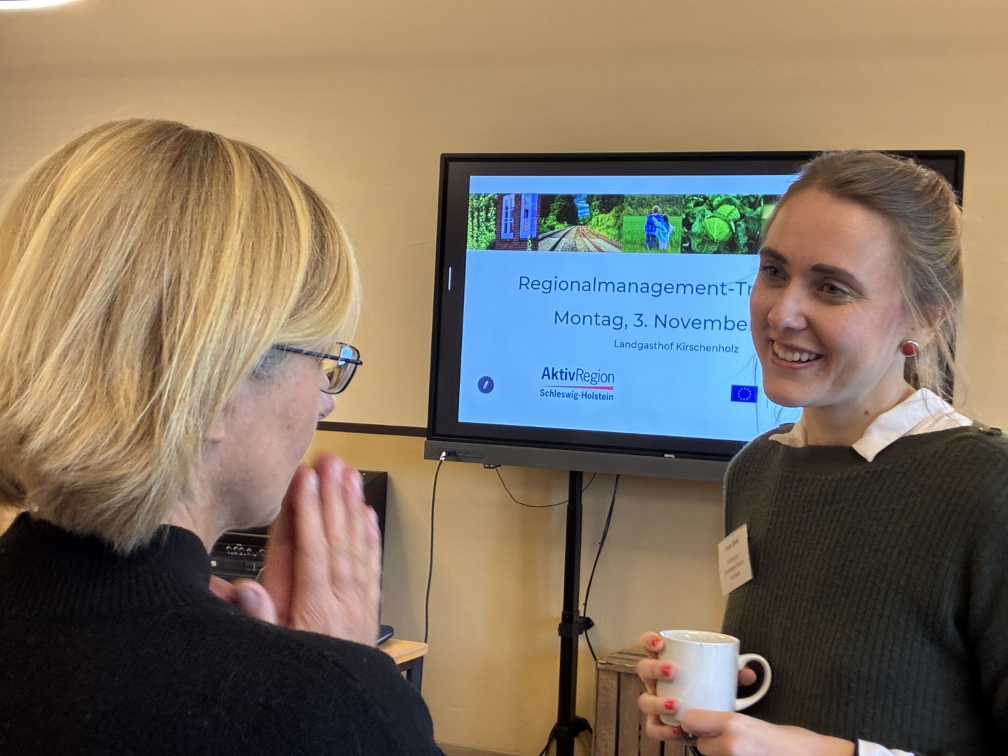 Zwei Frauen unterhalten sich, eine davon lächelt und hält eie Tasse in der Hand. Im Hintergund hängt ein großer Bildschirm an der Wand, auf dem steht: Regionalmanagement-Treffen