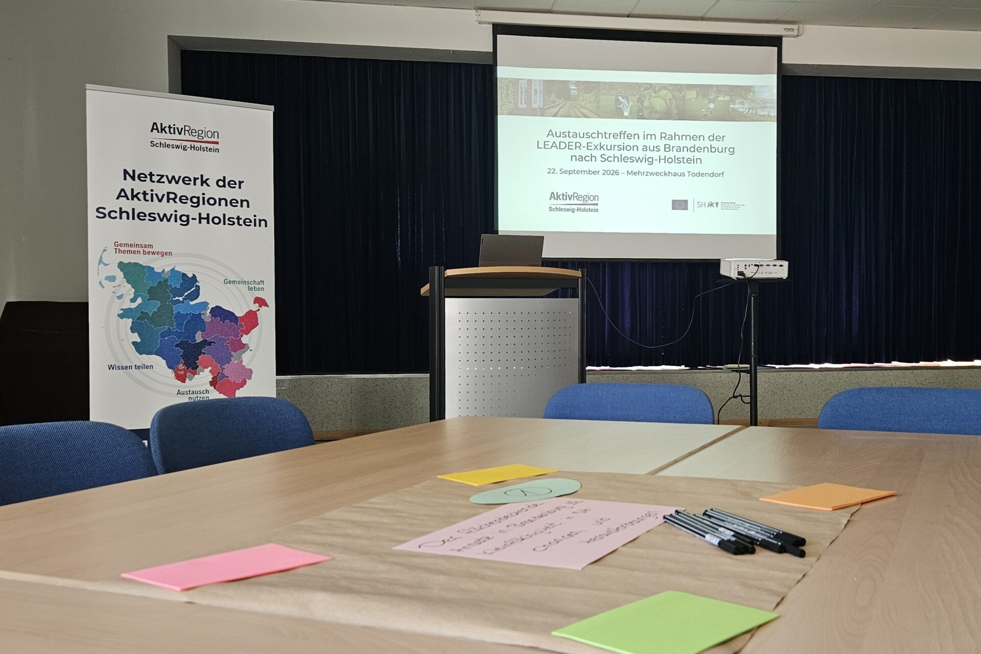 Seminarraum mit Konferenztisch, Post it's und Stiften. Im Hintergrund steht ein Rollup und eine Leinwand mit einer Präsentation der AktivRegion Schleswig-Holstein.